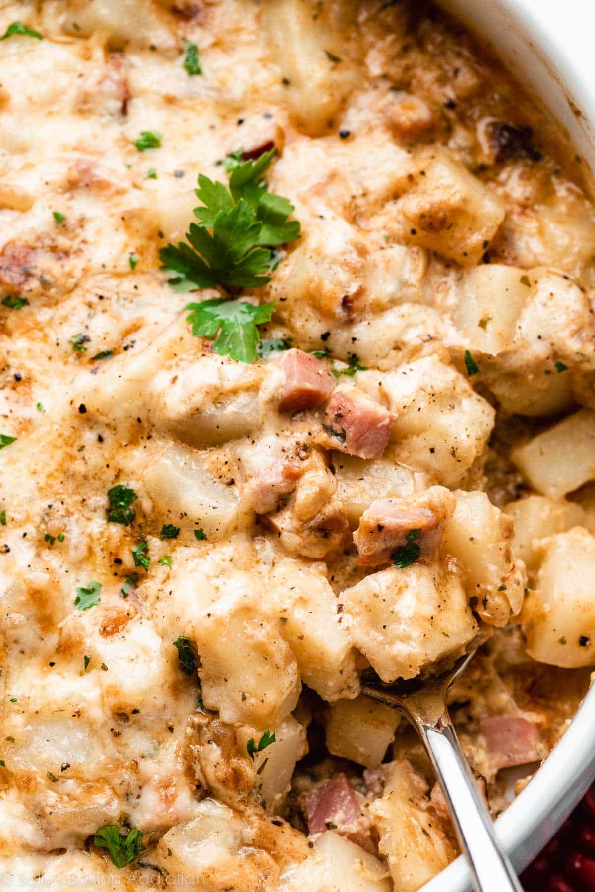 cheesy ham and potato casserole in white dish with serving spoon sitting in it.